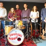 do zdjecia pozuje sześć młodych osób. muzyków. od lewej gitarzysta, skrzypaczka, prowadzacy zespół, perkusista, gitarzystka i basista. pozują w pomieszczeniu do prób.