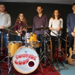 do zdjecia pozuje pięć młodych osób. muzyków. od lewej gitarzysta, skrzypaczka, perkusista, gitarzystka i basista. pozują w pomieszczeniu do prób.