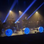 zdjecie z koncertu. meżczyzna na wiolonczeli, renata przemyk z małym bębnem, mężczyzna w koku na basie oraz gitarzysta. za nimi niebieskie światło, z przodu sceny 4 niebieskie lampy.