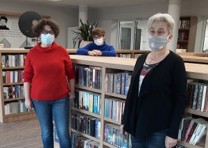 zdjęcie wykonane w pomieszczeniu bibliotecznym. do zdjecia pozują stojące przy regałach z książkami 3 bibliotekarki w maskach na twarzach