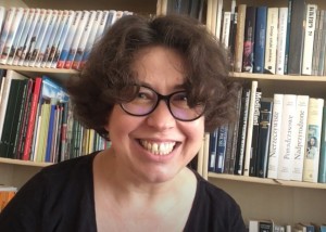na zdjęciu uśmiechnięta bibliotekarka w okularach siedząca na tle regału z książkami