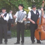 na zdjęciu 3 muzyków zespołu ludowego. grają na świeżym powietrzu, wszyscy elegancko ubrani i w kapeluszach. od lewej klarnecista, akordeonista i kontrabasista