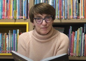 na zdjęciu uśmiechnięta bibliotekarka w okularach siedząca na tle regału z książkami