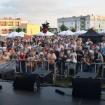 Zdjęcie zostało zrobione podczas koncertu, na pierwszym planie z lewej strony widzimy piosenkarza, w tle licznie zgromadzoną publiczność i budynki przedszkola i bloku w Opalenicy