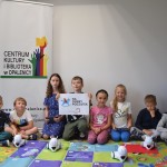 Zdjęcie zostało zrobione w pracowni Centrum Kultury i Biblioteka, na kolorowym fioletowo-zielonym dywanie siedzi ośmioro dzieci, czwarta od lewej - chłopiec trzyma kartkę, na której jest hasło Na dobry początek oraz logo w kształcie niebieskiego bujanego konia, przed dziećmi znajdują się cztery roboty, za dziećmi baner z logiem Centrum Kultury i Biblioteka w Opalenicy, z kolorowymi rękoma - zielona, niebieska, czerwona z żółtą książką oraz ręka żółta