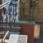 na zdjęciu na pierwszym planie tabliczka z kodem QR Opalenickiego Szlaku Historycznego, w tle budynek Ośrodka Pomocy Społecznej w Opalenicy, kamienne schody, białe plastikowe drzwi z dużymi szybami na mrze po prawej stronie Godło Polski, a także czerwona tabliczka z nazwą Ośrodek Pomocy Społecznej w Opalenicy, po prawej stronie doniczka z kwiatami