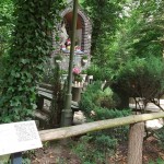 na zdjęciu na pierwszym planie tabliczka z kodem QR Opalenickiego Szlaku Historycznego,w tle kapliczka przy lesie w Opalenicy, a także zielone krzewy i drzewa oraz kawałek słupa od lampy