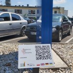 na zdjęciu na pierwszym planie tabliczka z kodem QR Opalenickiego Szlaku Historycznego, w tle samochody i budynek Cukrowni w Opalenicy, a także powierzchnia z jasnych kamieni