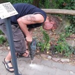 na zdjeciu młody mężczyzna w pozycji pochylonej, używający wiertarki, obok niego stoi tabliczka z kodem QR Opalenickiego Szlaku Historycznego