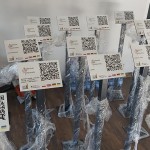 Na zdjęciu 17 tabliczek z kodem QR, słupki owinięte są w taśmy, po prawej stronie kawałek drzwi