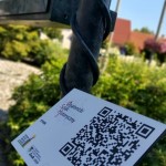 na zdjęciu tabliczka z kodem QR Opalenickiego Szlaku Historycznego, na tabliczce znajduą się flagi Polski, Wielkiej Brytanii, Niemiec i Ukrainy, a także loga Centrum Kultury i Biblioteki w Opalenicy oraz herb Opalenicy, w tle zielone krzaki znajdujące się na Rynku w Opalenicy