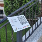 na zdjęciu na pierwszym planie tabliczka z kodem QR Opalenickiego Szlaku Historycznego, w tle kawałek poręczy w kolorze ciemnym przy schodach przy Urzędzie Miasta w Opalenicy, za poręczą stoi zielony krzew