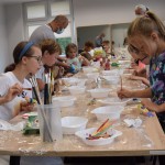 Zdjęcie zostało wykonane w pracowni Centrum Kultury i Biblioteka w Opalenicy, na środku zdjęcia znajduje się jasny stół owinięty folią, a na nim białe plastikowe miseczki z farbami i pędzlami, wokół stołu znajdują sie dzieci w kolorowych ubraniach