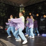 Zdjęcie zostało zrobione w plenerze - przed Muzeum w Sielinku. Na scenie tańczy grupa dziewczyn z Opalenica Dance Studio. Ubrane są w żółto fioletowe kurtki i jasno jeansowe spodnie.