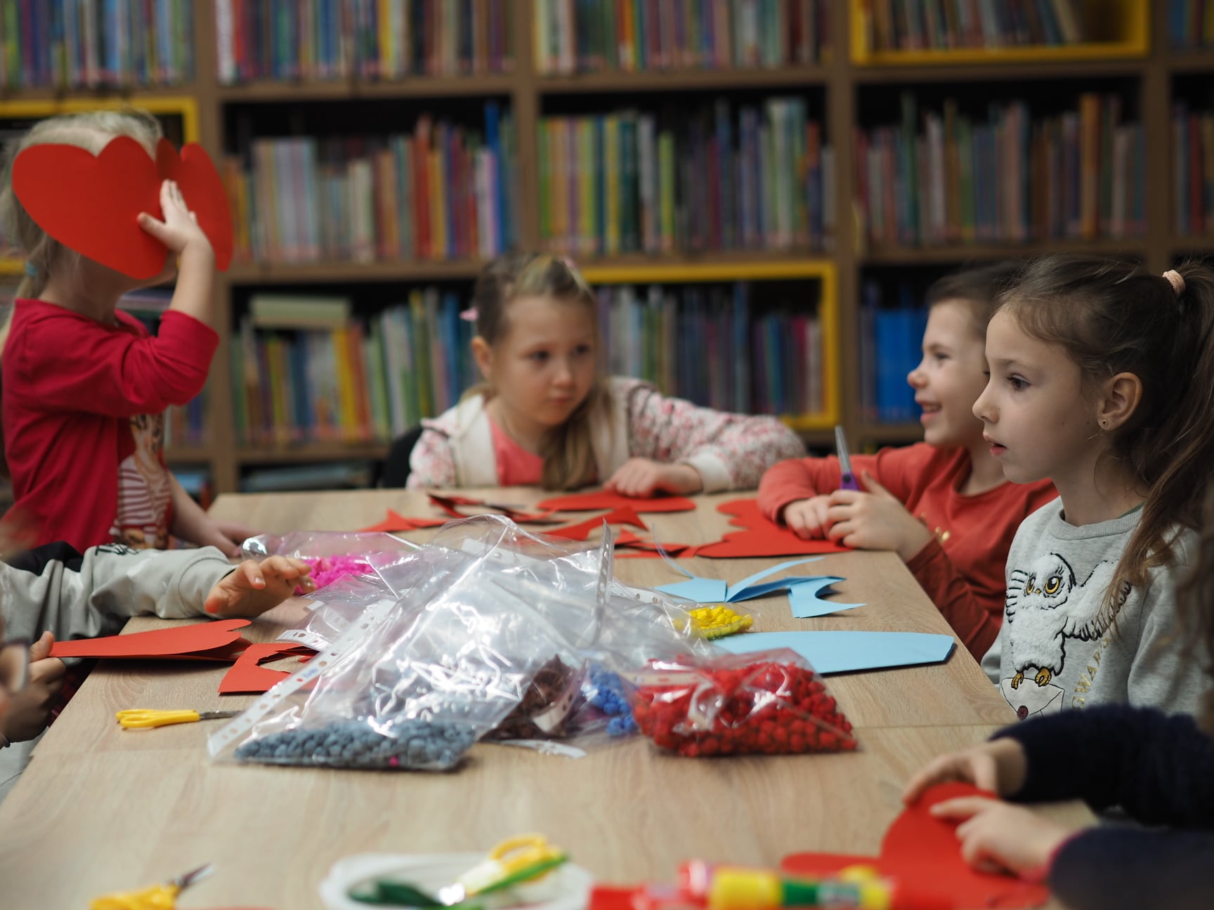 Zdjęcie zostało zrobione w bibliotece podczas zajęć plastycznych Dzieci siedzą wokół stołu. Na stole są różne papiery. i ozdoby.