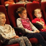 Zdjęcie przedstawia publiczność podczas teatrzyku kukiełkowego. Dzieci siedzą na karminowych fotelach.