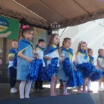grupa dzieci występująca na scenie, ubrana na niebiesko z żółtymi elementami, w rękach trzymają niebieskie pompony