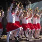 dziewczynki na scenie ubrane w czerwone spódniczki i białe bluzki, trzymają ręce do góry i trzymają wianki z kwiatów