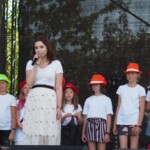 na zdjęciu młoda dziewczyna na scenie z mikrofonem w ręce, za nią stoją w rzędzie dzieci ubrane w czerwone i zielone kapelusze