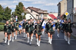 mażoretki ubrane w ciemno zielone stroje idą ulicą, a za nimi grająca orkiestra