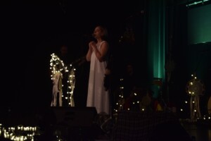 wokalistka zespołu stoi na scenie w klimatycznej scenerii, zgaszone są światła, świecą tylko małe lampki ozdobne