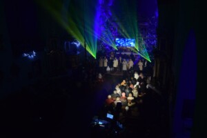 zdjęcie przedstawia publiczność oraz opalenicką grupę wokalną podczas występu w kościele, otoczenie jest ciemne, światła padają tylko na artystów oraz część publiczności