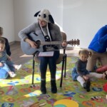 na zdjęciu prowadząca zajęcia z gitarą, dookoła na dywanie siedzą rodzice z dziećmi