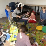na zdjęciu prowadząca zajęcia z gitarą, dookoła na dywanie siedzą rodzice z dziećmi