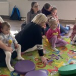 na zdjęciu prowadząca zajęcia z gitarą, dookoła na dywanie siedzą rodzice z dziećmi