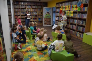 grupa przedszkolaków siedzi na dywanie w bibliotece, przed nimi wykład prowadzi Pani Elżbieta Adamczewska, która trzyma w rękach książke