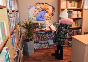 Zdjecie wykonane w pomieszczeniu bibliotecznym. na pierwszym planie obrócona tyłem do obiektywu dziewczynka w zimowym ubraniu czyta książkę. dookoła nije stoją regały z książkami