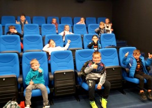 Zdjęcie zostało wykonane w sali kinowej Kina za Rogiem w Centrum Kultury i Biblioteka w Opalenicy, na pierwszym planie widzimy niebieskie krzesła kinowe, w tle na krzesłach siedzi 12 młodych osób