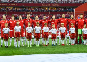 zdjecie przedstawia piłkarzy polskiej reprezentacji piłki nożnej stojących na boisku. Przed piłkarzami stoją dzieci w strojach piłkarskich