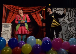 Zdjęcie zostało zrobione w sali widowiskowej w Centrum Kultury i Biblioteka w Opalenicy, na scenie widzimy dwójkę aktorów - po lewej kobietę z grymasem na twarzy i w różowej sukience z falbanami, po prawej aktora w okularach grającego pchłę, na dole leżą kolorowe balony, w tle widać zdjęcie kurtyny