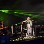 Zdjęcie zostało wykonane podczas plenerowego koncertu, na scenie widzimy trójkę muzków - wokalistka ubrana jest w białą bluzkę i spodnie, na scenie leży sznur lampek o silnym natężeniu światła