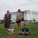 Zdjęcie zostało wykonane na dworze, na pierwszym planie widzimy siedzące na trawie dwójkę dzieci - po lewej dziewczynka po prawej chłopiec, za nimi stoją dwie dorosłe kobiety, w tle widać budynek bloku i bramki do gry w piłkę nożną
