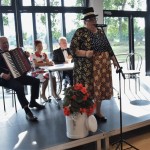 Zdjęcie zostało zrobione w Sali Wiejskiej w Urbanowie. Na scenie znajdują się cztery osoby. Jedna z nich stoi - to kobieta ubrana w granatową bluzkę w kropki i spódnicę w kolorowy wzór. Na głowie ma cylinder. Za nią siedzą przy stole trzy osoby. Od lewej pan ubrany w garnitur, trzymający akordeon, kobieta ubrana w biała sukienkę w czerwone kwiaty i pan ubrany w garnitur z muszką. Za nimi są wielkie okna, przez które wpada światło.