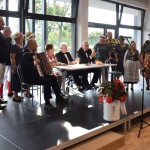 Zdjęcie zostało zrobione w Sali Wiejskiej w Urbanowie. Na scenie znajduje się 11 osób. Są różnokolorowo ubrani. Cztery z ich siedzą przy stole. Za nimi są wielkie okna, przez które wpada światło.