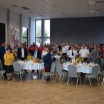 Zdjęcie zostało zrobione w Sali Wiejskiej w Urbanowie. Zdjęcie przedstawia zgromadzoną publiczność, która stoi przy okrągłych stolikach.
