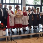 Zdjęcie zostało zrobione w Sali Wiejskiej w Urbanowie. Na scenie stoi 11 osób. Trzy kobiety pochodzą z zespołu Rudniczoki i mają czerwone suknie z białymi fartuszkami. Reszta osób ubrana jest w ciemne ubrania.Za nimi są wielkie okna, przez które wpada światło.