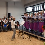Zdjęcie zostało wykonane w Sali Wiejskiej w Urbanowie. Na scenie stoi chór. Kobiety ubrane są w fioletowe spódnice i wzorzyste bluzki. Pan stojący za nimi ma fioletową kamizelkę i białą koszulę. Przed nimi stoi dyrygent, trzymający akordeon, ubrany jest w biały sweter i szare spodnie. Obok ich siedzi zespół Wiaruchna, ubrany w ciemne kolory. Panie mają białe kołnierzyki.