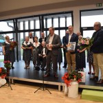 Zdjęcie zostało zrobione w Sali Wiejskiej w Urbanowie. Przy mikrofonie stoi Burmistrz Opalenicy - Tomasz Szulc, obok niego Grzegorz Mądroszyk - Promocja UM w Opalenicy i Dyrektor CKiB w Opalenicy Krzysztof Sadowski, który trzyma różowo-zielone kwiaty. Za nimi stoi zespół Wiaruchna.