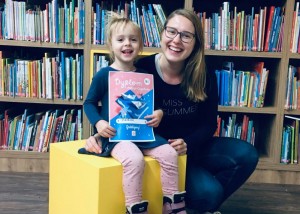 Zdjęcie zostało zrobione w bibliotece ckib w Opalenicy. Przed regałem z książkami na żółtym klocku siedzi mała dziewczyka trzymająca dyplom, obok nie kuca uśmiechnięta mama. Kobieta ubrana jest w ciemne kolory. Na twarzy ma okulary.