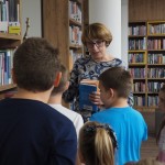 Zdjęcie zostało zrobione w Bibliotece w CKiB. Dzieci na pierwszym planie stoją odwrócone tyłem do aparatu i oglądają regały. Kobieta w średnim wieku tłumaczy im. W ręce trzyma książki. Jest odwrócona przodem do kamery.