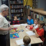 Zdjęcie zostało zrobione w bibliotece CKiB w Opalenicy. Na zdjęciu wśród regałów podstawiony jest stół, na którym leżą kolorowe kredki i białe kartki papieru. Wokół stołu siedzi pięciu chłopców. Przed nimi stoi starsza kobieta tłumacząca zasady zabawy.