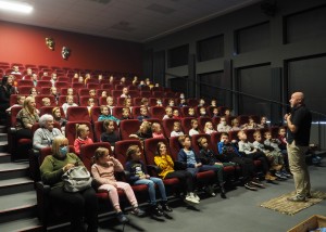 Zdjęcie zostało zrobione w sali widowiskowej w CKiB w Opalenicy. Zdjęcie przedstawia siedzące dzieci w sali widowiskowej na karminowych fotelach. Przed nimi stoi pan w średnim wieku, ubrany w czarną bluzkę i spodnie w kolorze khaki.