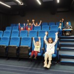 Na zdjęciu dzieci siedzące na niebieskich krzesłach w kinie. Dzieci mają podniesione ręce do góry.