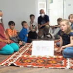 dzieci i prowadzący siedzą na podłodze wokół czerwonego ornamentalnego dywanu
